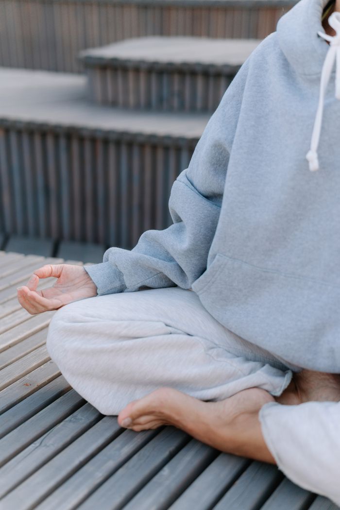 Person meditating in a gray hoodie and white pants, embracing a healthy lifestyle outdoors.